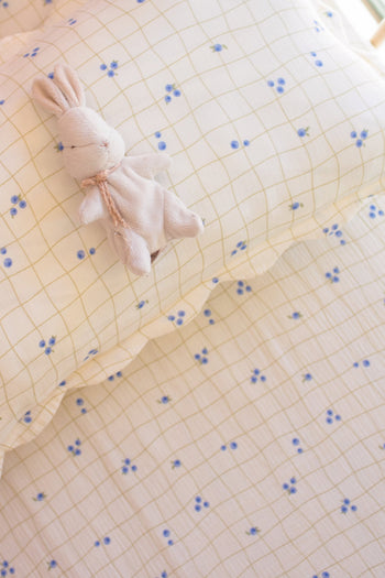 A small, plush bunny toy rests on a Soft Spot Soft Petal Rectangle Cushion Cover featuring a yellow grid and blue flowers, matching the bedding underneath.