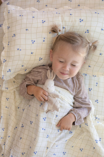 A young child with blonde pigtails lies in bed on a Soft Spot Soft Petal Cushion Cover, covered with a blanket and holding a soft toy bunny while smiling gently.
