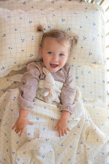 A smiling toddler with light hair in pigtails lies in bed holding a stuffed bunny, resting on Soft Spot Soft Petal Cushion Covers Rectangle, Set of 2.