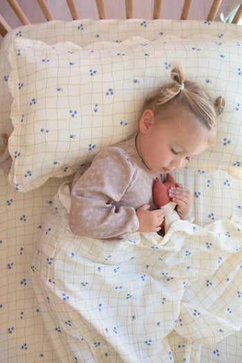 A young child with blonde hair in two buns sleeps in a crib, cuddling a stuffed toy, covered by Soft Spot’s Soft Petal Cushion Covers Rectangle (set of 2) featuring a cream grid and blue flower pattern.