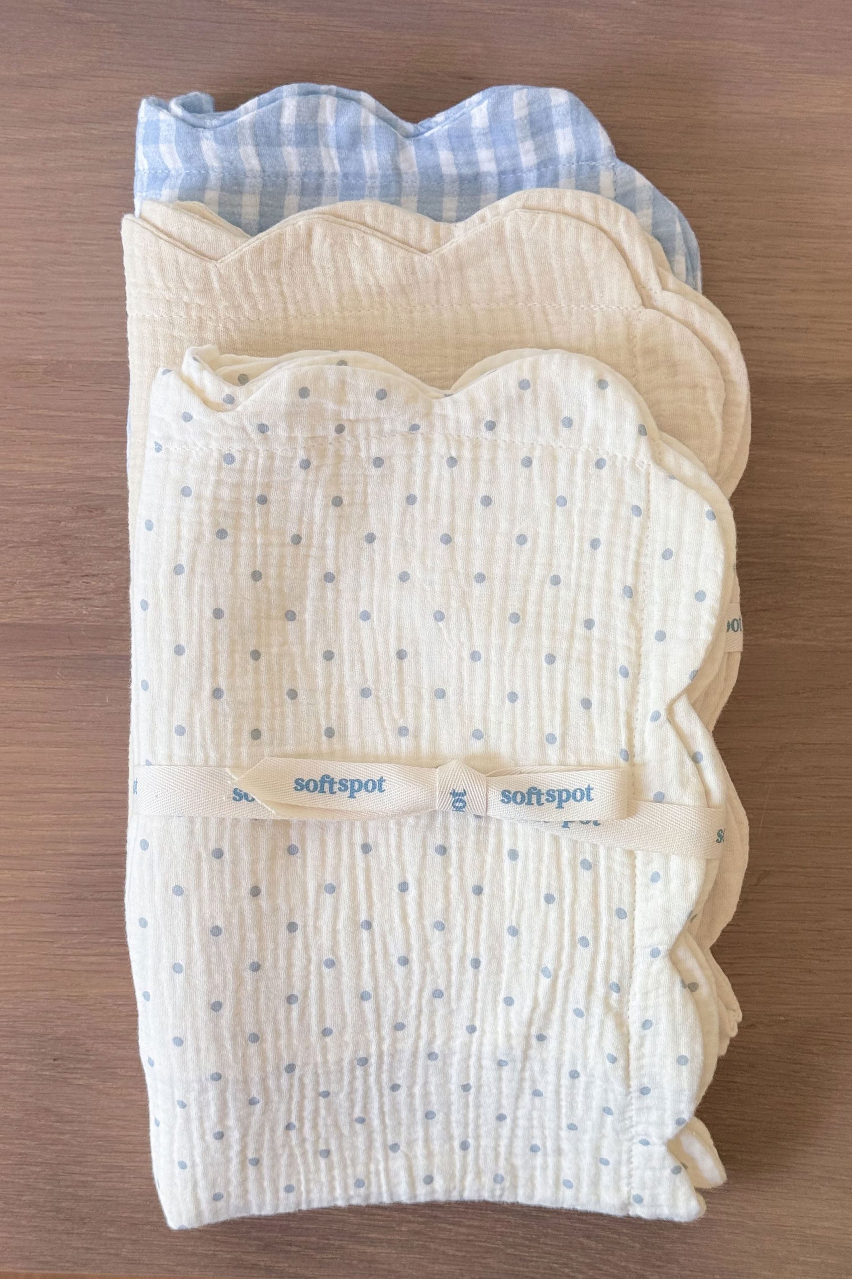 A set of two Soft Spot Soft Petal Cushion Covers Rectangle are neatly tied together with a branded ribbon and arranged on a wooden surface, creating a charming display reminiscent of scallop-edged muslin blankets in cream and blue gingham.