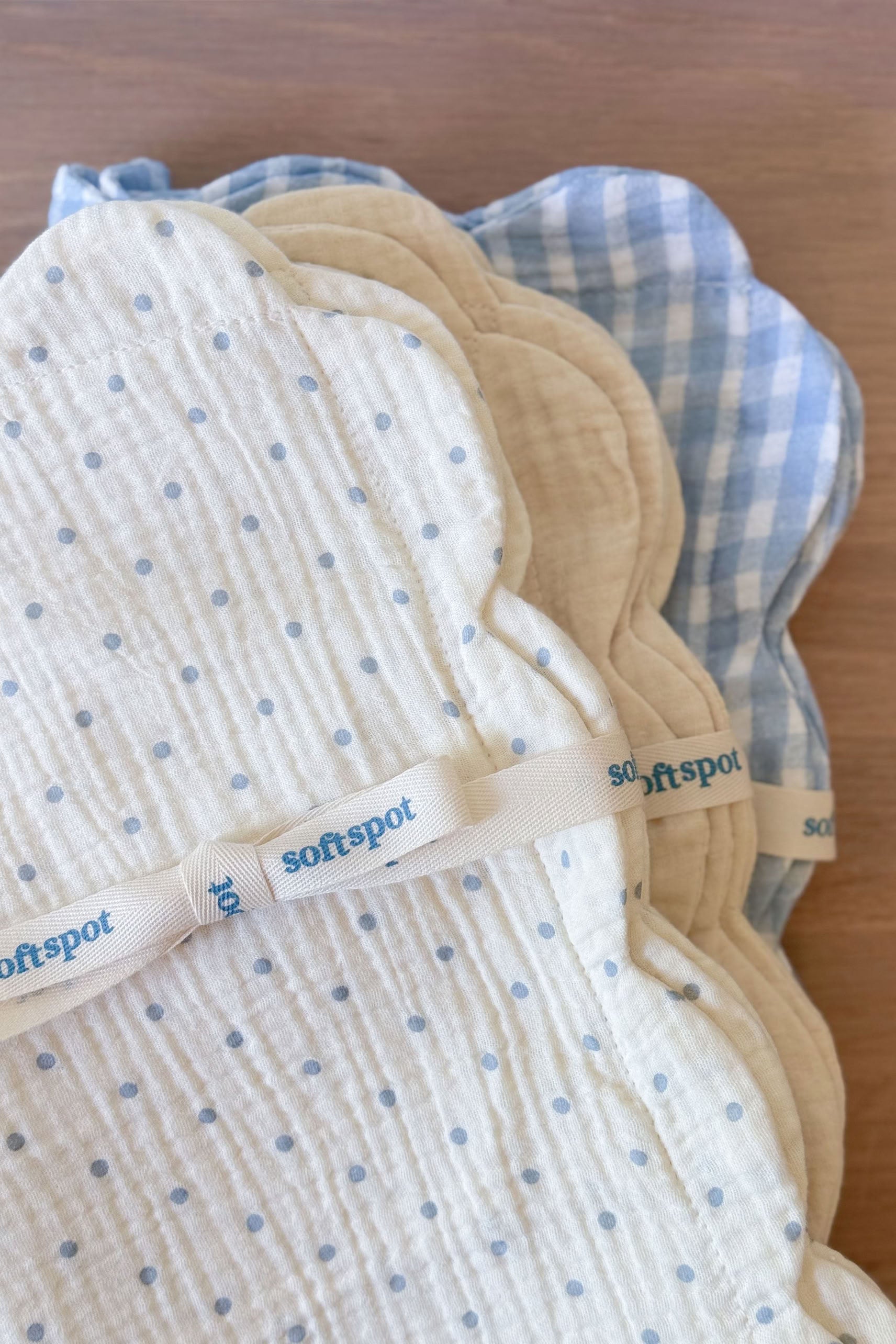A set of two Soft Spot Soft Petal Cushion Covers Rectangle—featuring a stack of cloud-shaped, quilted cream pads adorned with blue dots and blue gingham patterns, tied together with a ribbon labeled "Soft Spot"—rests on a wooden surface.