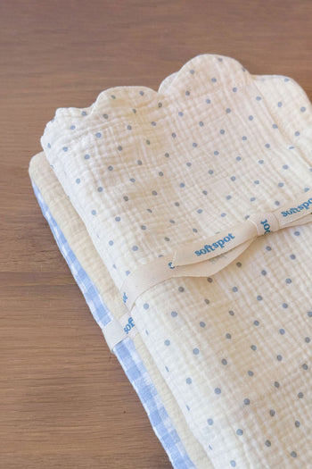 Two folded baby blankets—one white with blue polka dots, the other in a blue and white check—rest on a wooden surface, neatly tied together with a cream Soft Spot ribbon. Just like our Soft Spot Soft Petal Cushion Covers Rectangle, Set of 2, these blankets are designed to perfectly complement your nursery décor.
