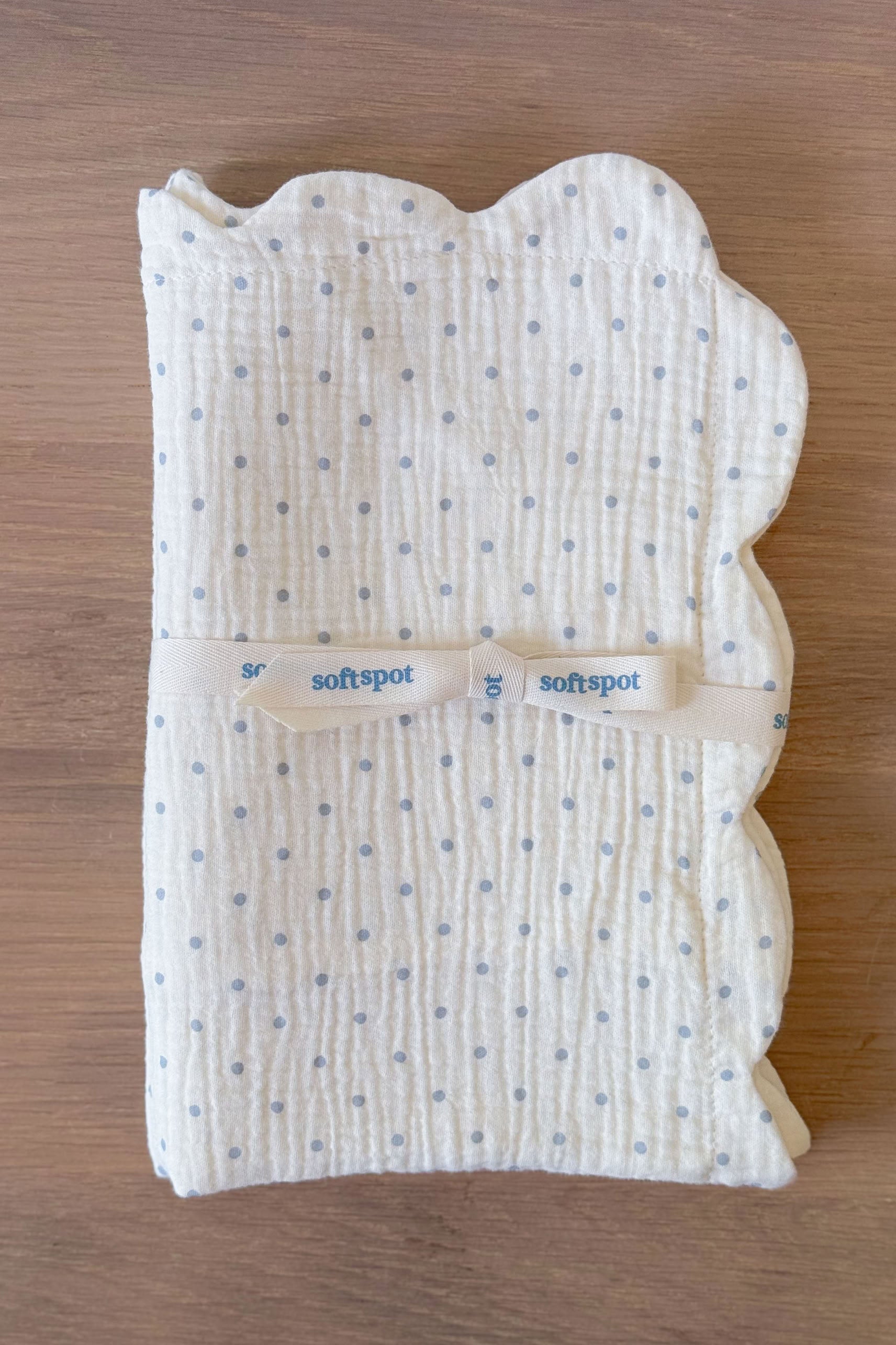 A set of two Soft Spot Soft Petal Cushion Covers in a rectangular shape, featuring scalloped petal-like edges, crafted from white muslin with small blue polka dots and finished with a cream ribbon labeled "softspot," displayed on a wooden surface.