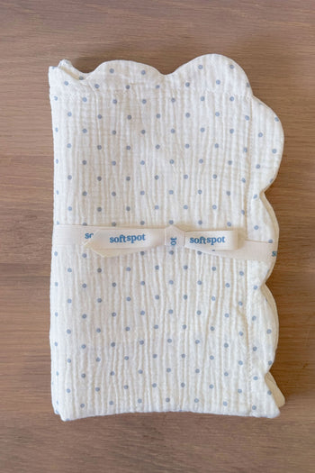 A set of two Soft Spot Soft Petal Cushion Covers in a rectangular shape, featuring scalloped petal-like edges, crafted from white muslin with small blue polka dots and finished with a cream ribbon labeled "softspot," displayed on a wooden surface.