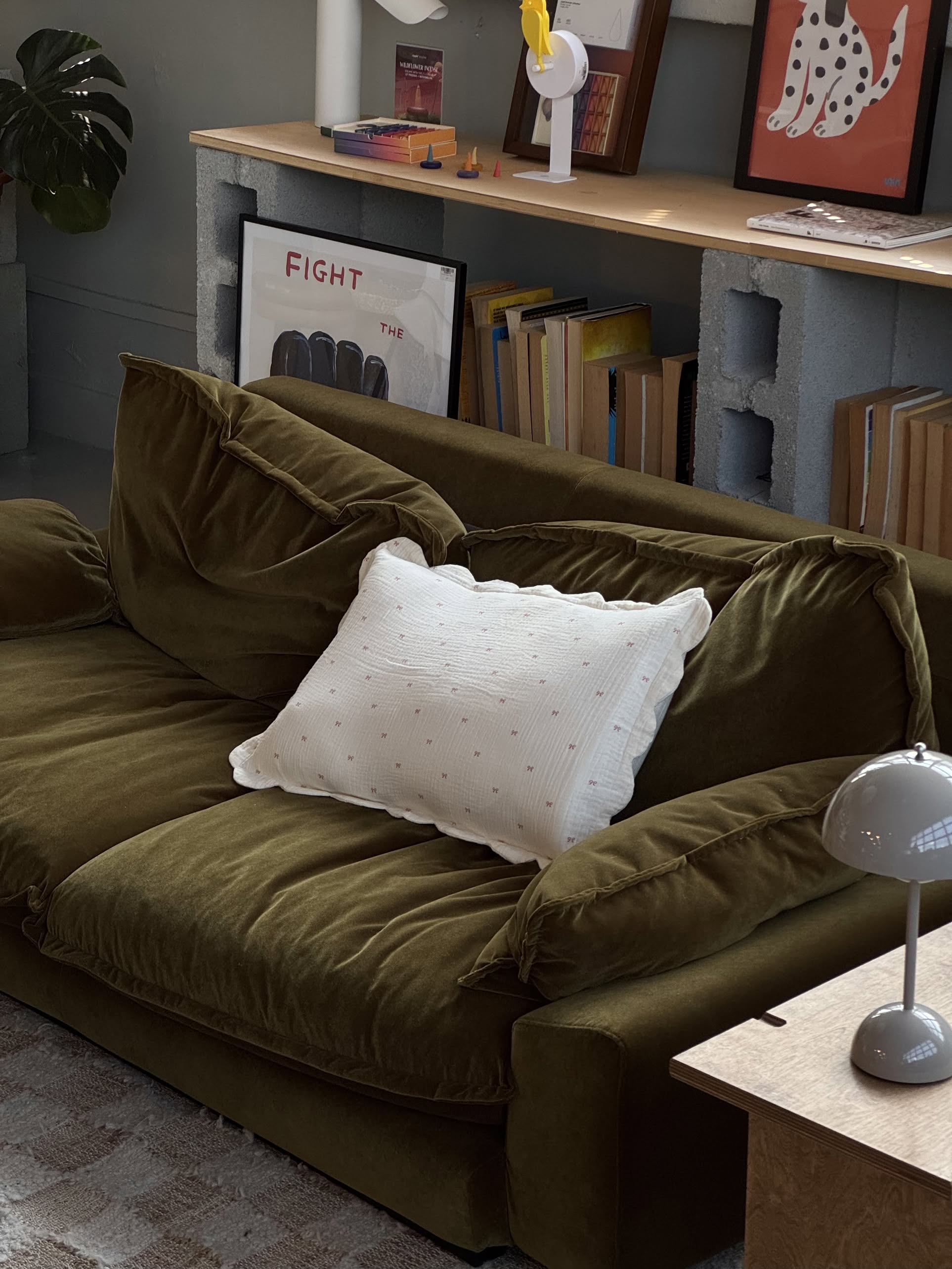 A green velvet sofa with Soft Spot Soft Petal Cushion Covers Rectangle (set of 2) and a white pillow sits in a cozy room. Behind it, books and framed art rest on a cinder block shelf, with a gray lamp and wooden table in the foreground.