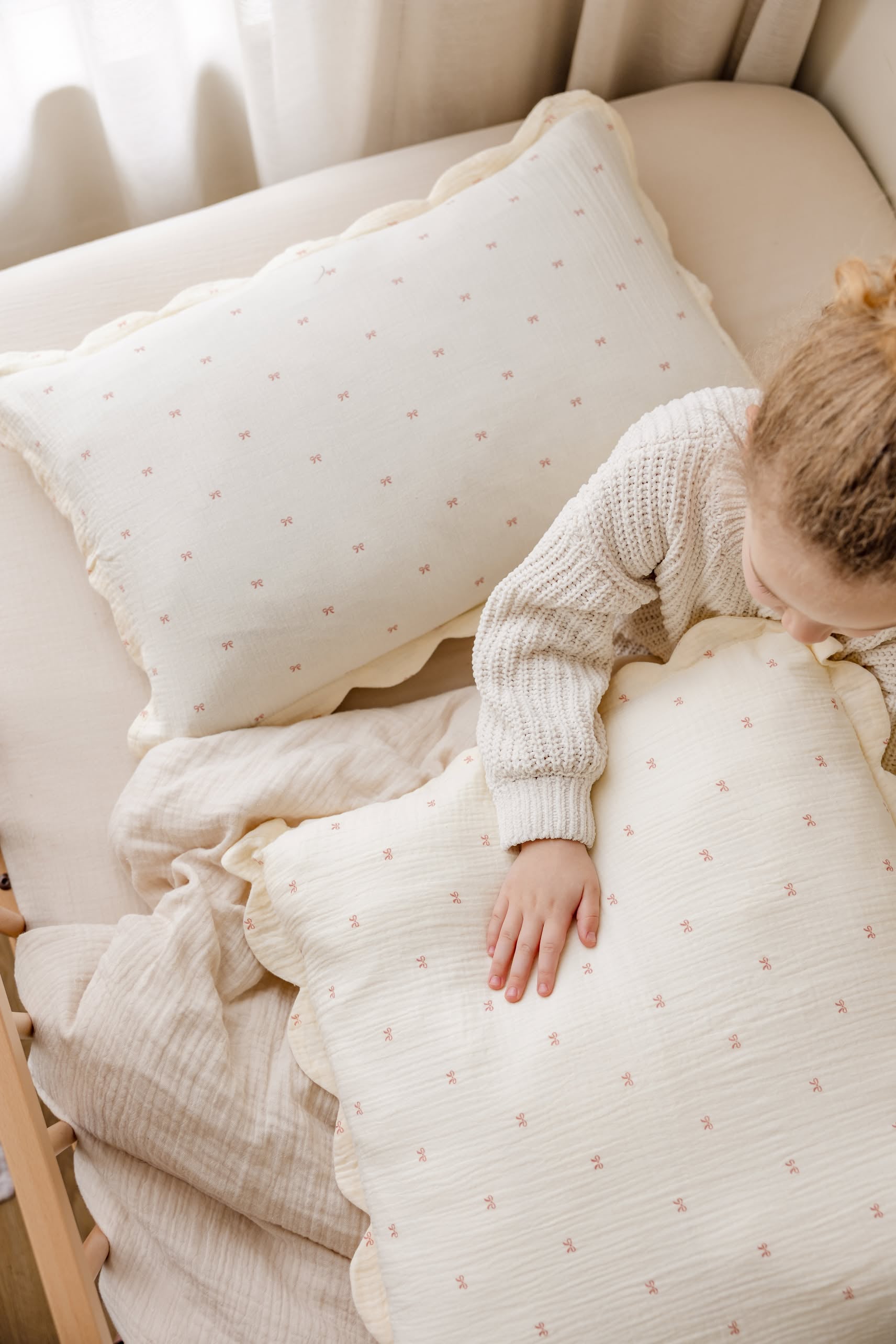 A young child wearing a beige knit sweater rests on cream bedding, gently touching one of the Soft Spot Soft Petal Cushion Covers Rectangle (Set of 2) by Soft Spot. Patterned pillows and soft natural light complete the cozy scene.