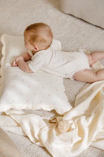A baby in a cream onesie lies on a soft carpet, resting on Soft Spot Soft Petal Cushion Covers Rectangle, Set of 2 by Soft Spot. A cream blanket and small woven rattle are nearby.