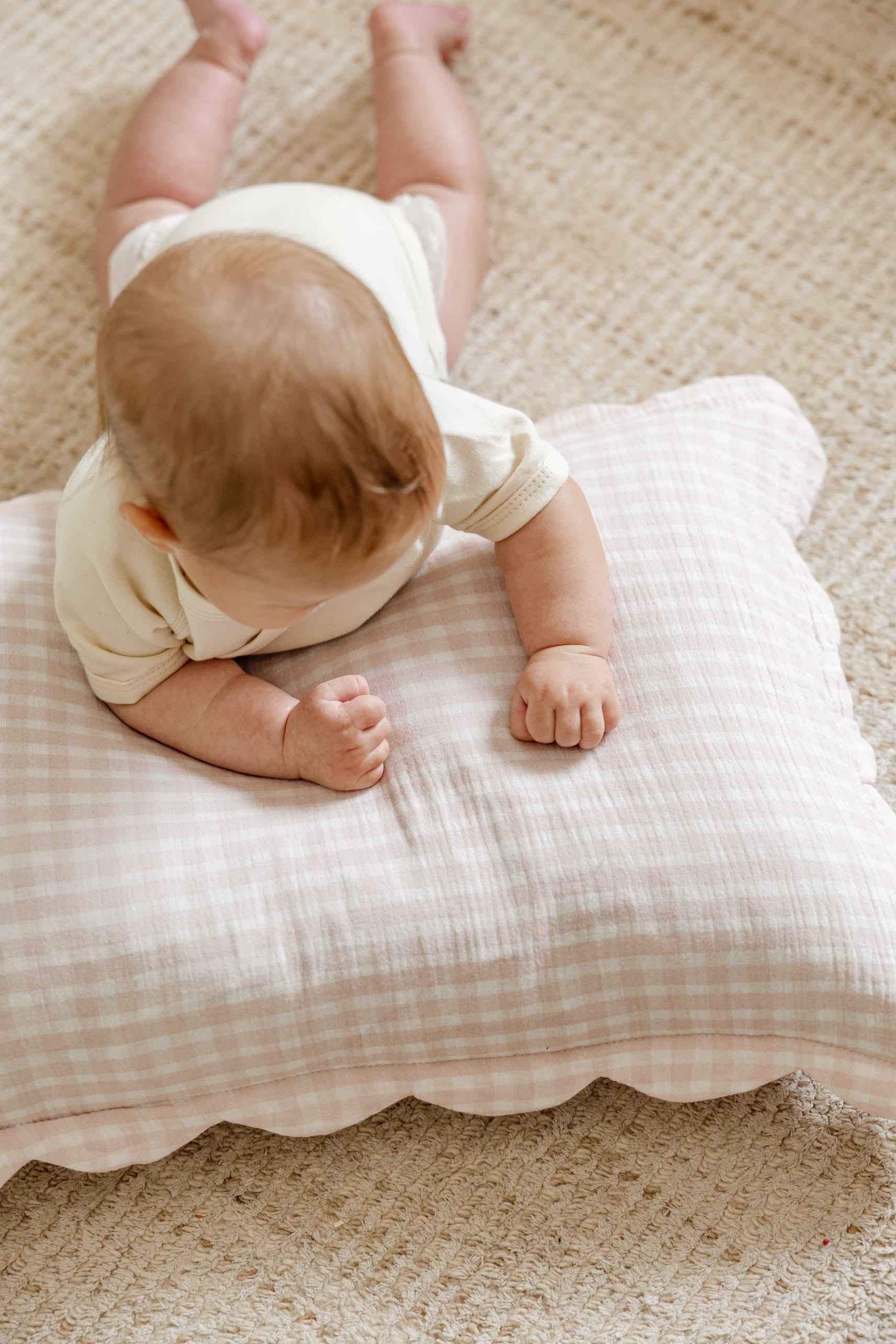 A baby rests on their stomach atop a textured beige rug, supported by Soft Spot Soft Petal Cushion Covers Rectangle, Set of 2, in light pink and white gingham, with their head turned down and arms on the cushions.