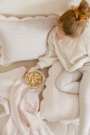 A young girl with curly hair in a bun sits on a bed with pink plaid bedding, holding popcorn. She wears a beige sweater and light leggings atop Soft Spot Soft Petal Cushion Covers Rectangle, Set of 2 by Soft Spot.