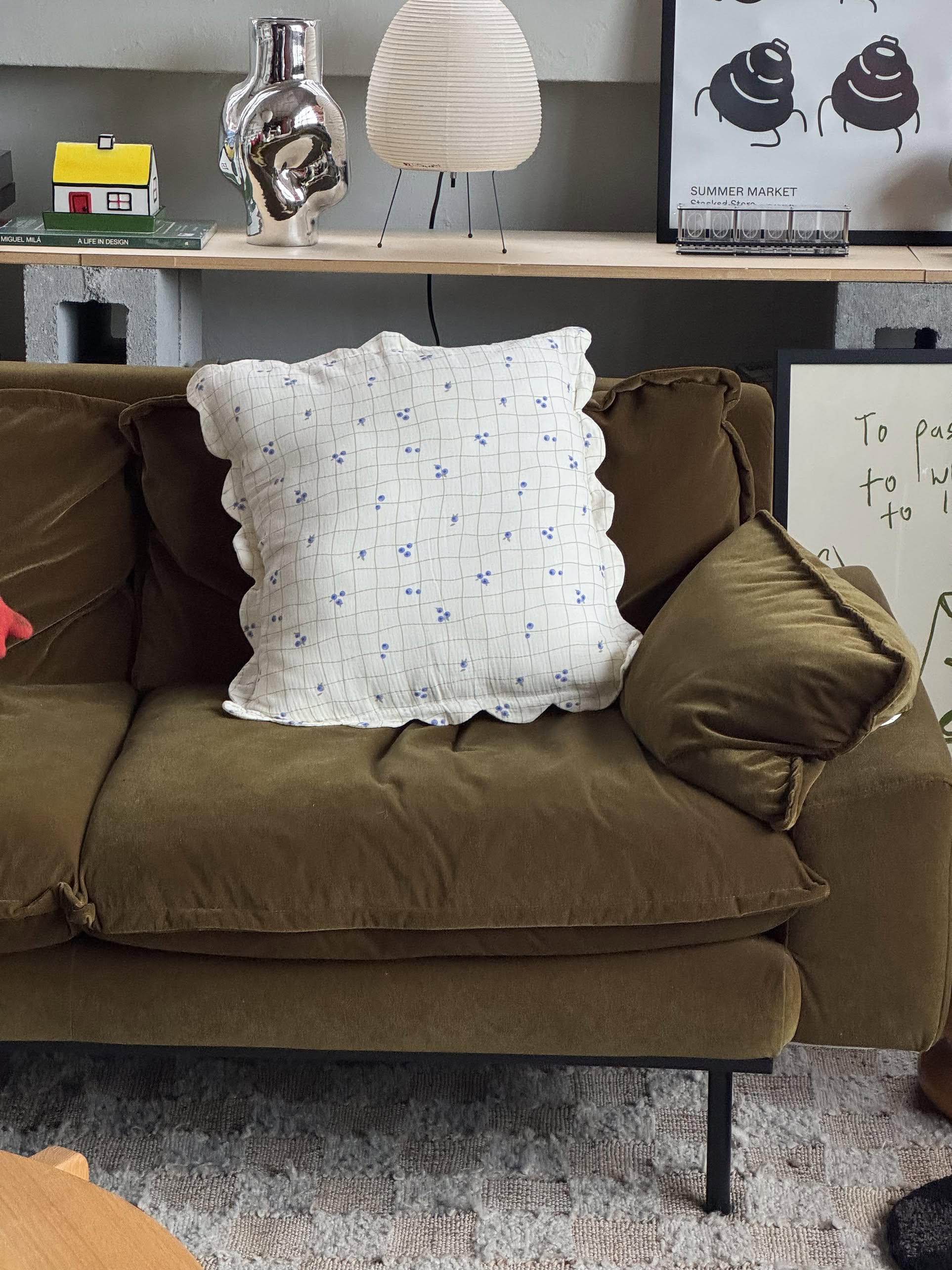 A large olive green couch with Soft Spot Soft Petal Cushion Covers, Set of 2 by Soft Spot, featuring a blue grid and small blue flowers, sits in a modern living room with decor and framed art on the shelf behind.