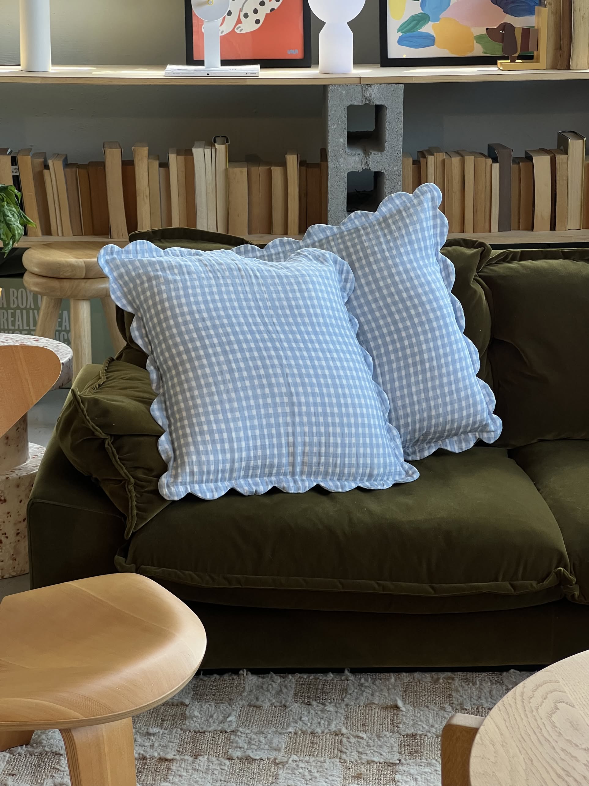 A small brown plush bear sits by a blue and white gingham pillow with scalloped edges, featuring Soft Spot Soft Petal Cushion Covers, Set of 2, on a wooden surface against a plain light-colored wall.