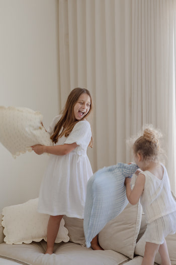 Two young girls laugh and toss Soft Spot Soft Petal Cushion Covers, Set of 2, made from light-colored fabric, on a beige sofa in a bright room with cream-colored curtains.