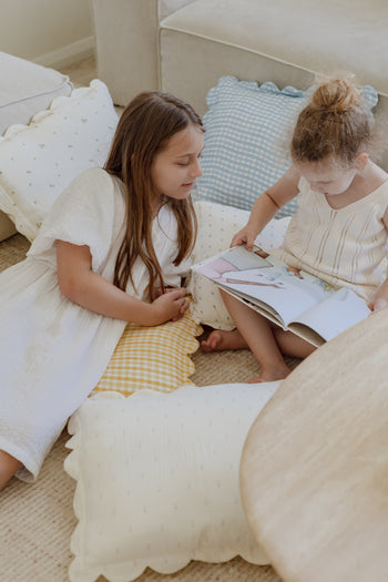 Two young girls relax on Soft Spot Soft Petal Cushion Covers, Set of 2, reading a picture book together. They are surrounded by Soft Spot’s soft, patterned pillows in a cozy, light-filled room.