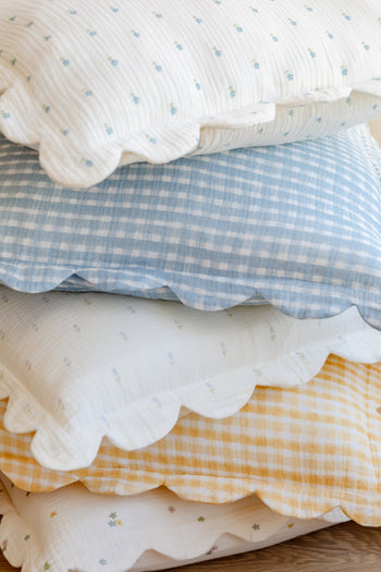 A stack of two Soft Spot Soft Petal Cushion Covers, featuring pastel florals and gingham patterns, is neatly arranged on a wooden surface.