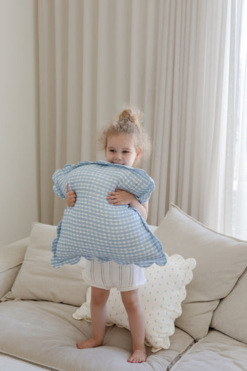 A young child with tied-up curly hair smiles on a beige couch, holding a blue gingham cloud pillow. The cozy scene features Soft Spots Soft Petal Cushion Covers (Set of 2) and light curtains by the window in the background.