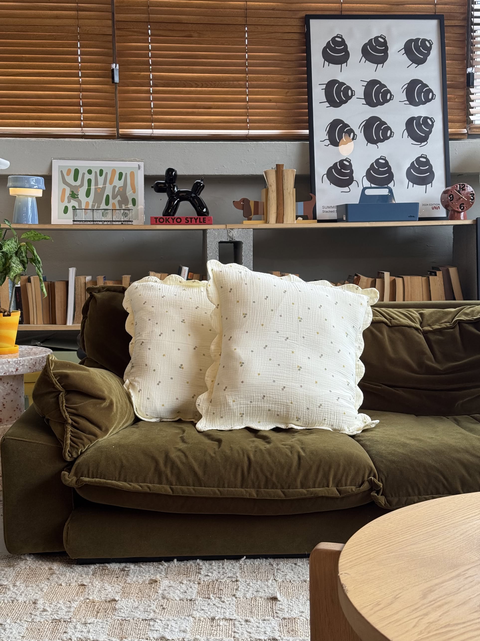 A cozy green velvet sofa with two Soft Spot Soft Petal Cushion Covers sits in front of a bookshelf filled with books, art prints, and decor. Wooden blinds cover the window above.