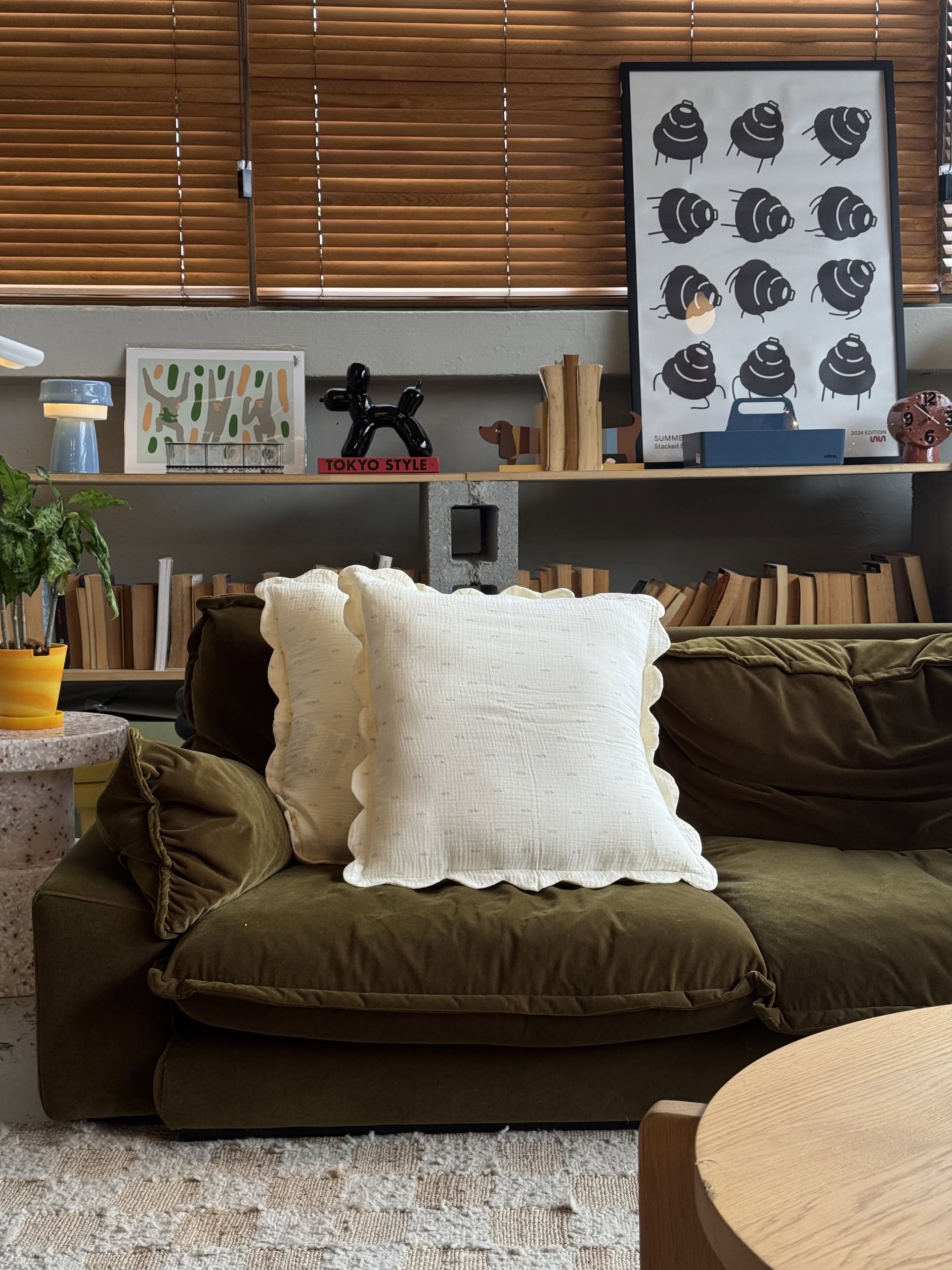 A cozy green sofa with two Soft Spot Soft Petal Cushion Covers sits in a stylish living room. Behind it, shelves display books, plants, and modern art prints; wooden blinds cover the window above.