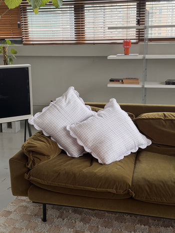 A modern living room with an olive green sofa features two Soft Spot Soft Petal Cushion Covers. Behind the sofa is a shelf with books, a red glass, and a large window with blinds. A leafy plant and TV complete the scene.