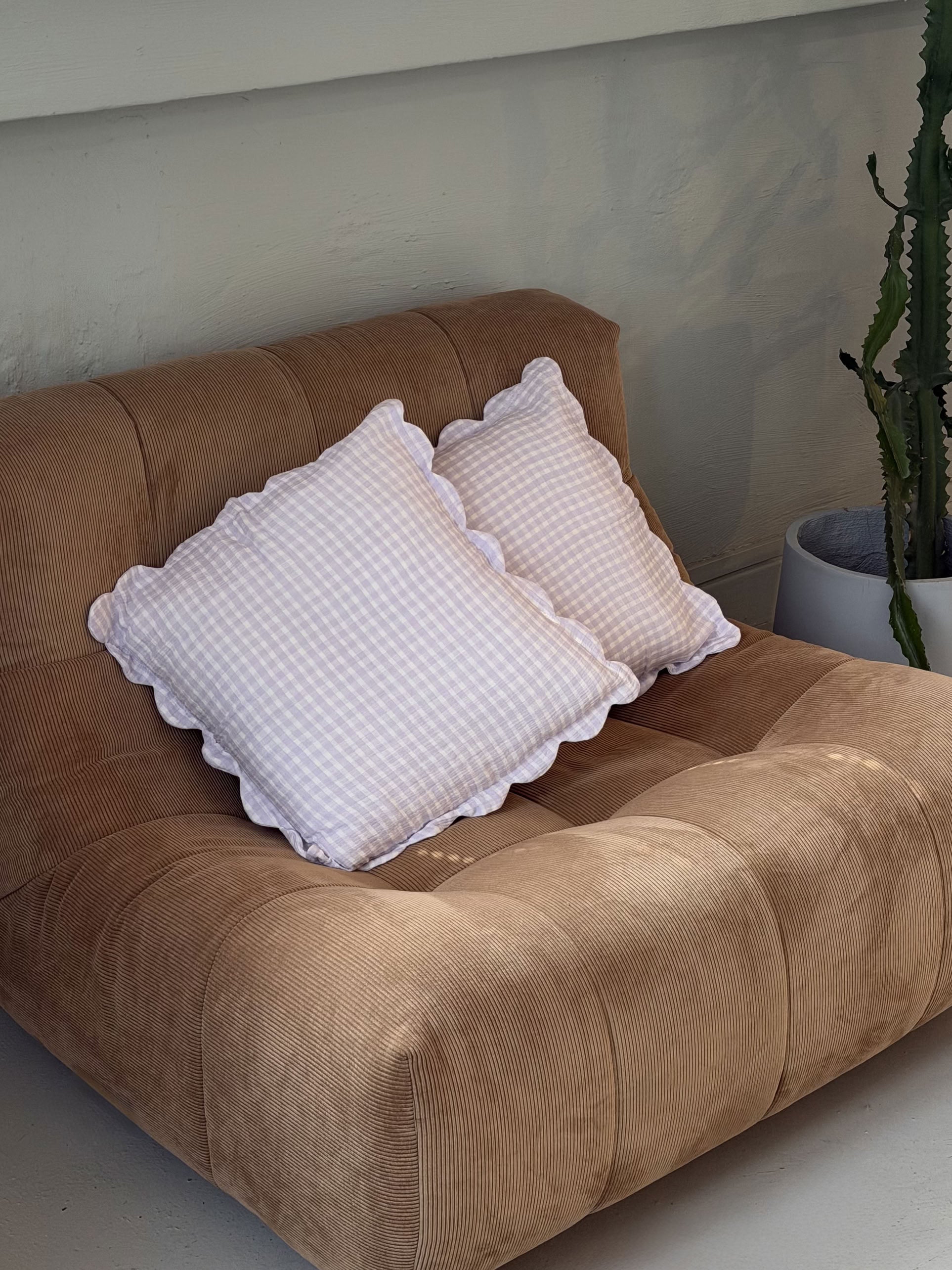 A plush brown corduroy sofa with two Soft Spot Soft Petal Cushion Covers by Soft Spot sits beside a large green cactus in a white planter against a light-colored wall.