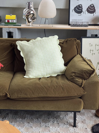 A brown velvet sofa with a Soft Spot Soft Petal Cushion Cover in light green and white checks sits in a cozy living room. Behind it are decorative items like a lamp, art prints, and a small yellow house model on a wooden shelf.