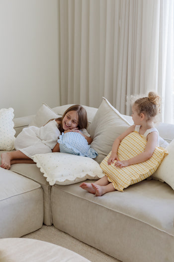 Two young girls smile on a light-colored sectional, holding Soft Spot Soft Petal Cushion Covers (Set of 2). Soft lighting and sheer curtains complete the cozy, relaxed atmosphere.