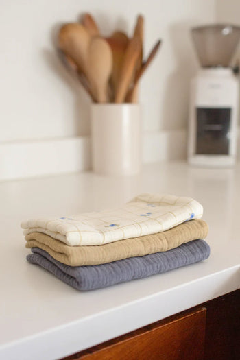The Soft Spot Soft Tea Towels (Set of 3) by Soft Spot, in white, tan, and gray, are stacked on a countertop. A container with wooden utensils and a coffee grinder sit nearby—versatile towels ideal for any kitchen.