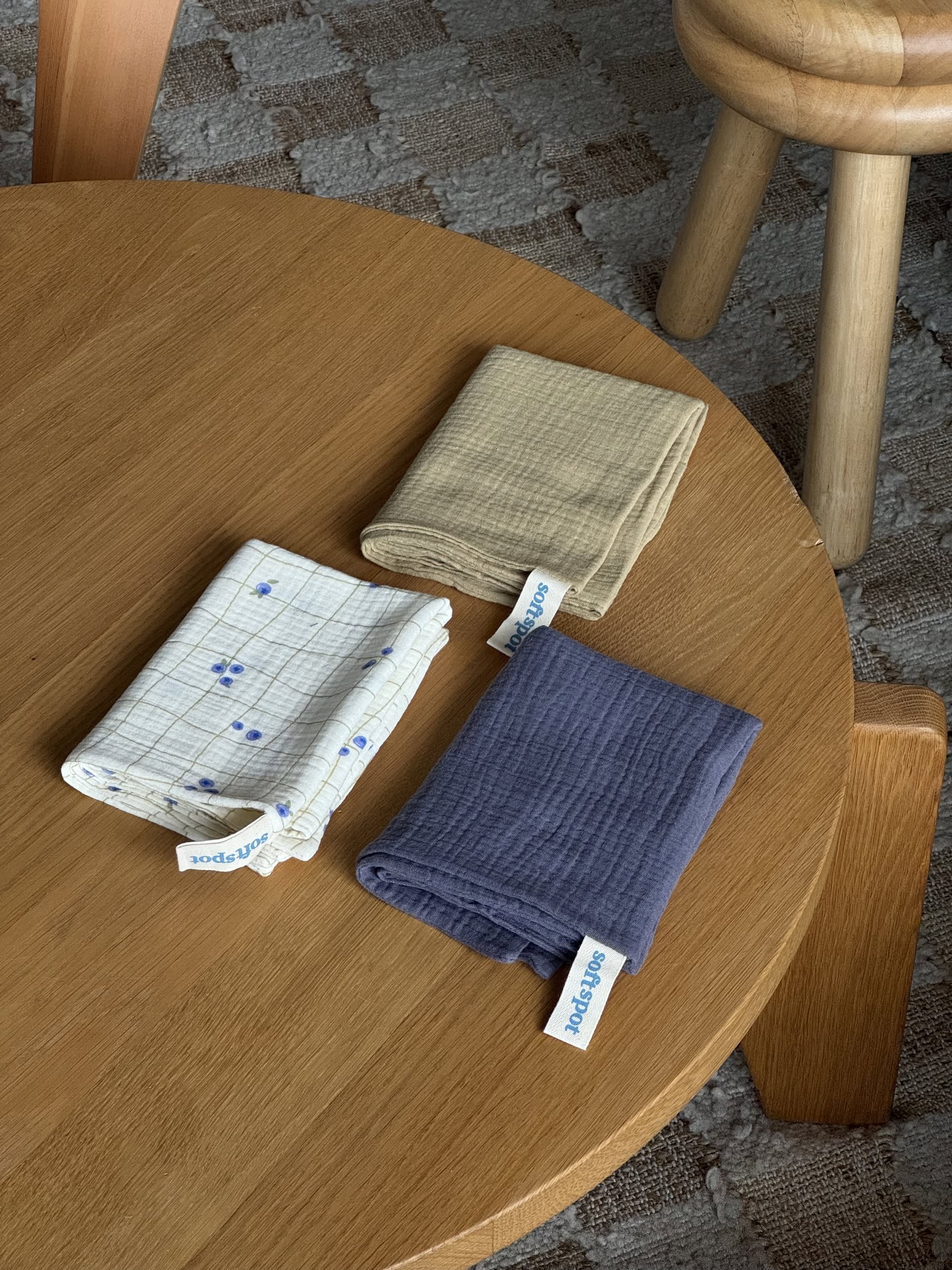 The Soft Spot Soft Tea Towels (Set of 3) by Soft Spot—patterned white, solid beige, and dark blue—are neatly folded on a round wooden table, with a wooden stool and textured carpet in the background.