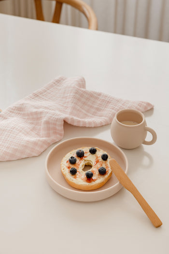 A bagel with cream cheese and blueberries sits on a plate beside coffee and a Soft Spot Soft Tea Towel by Soft Spot, all arranged on a white table.