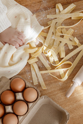 A child’s hand rests on a Soft Spot Soft Tea Towel (Set of 3) by Soft Spot beside fresh pasta strips on wood, with a carton of brown eggs, a small rolling pin, and flour scattered across the table.