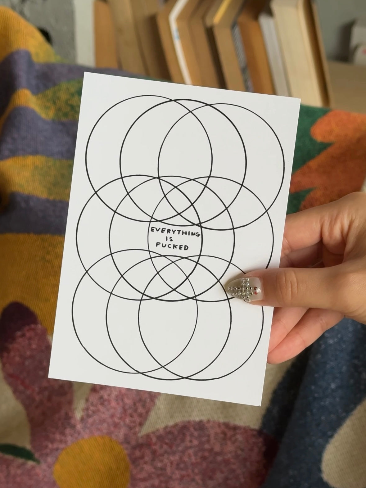 A hand with decorated nails holds the Stacked Store Everything Is Fucked Gift Card (Physical) by David Shrigley, displaying overlapping black circles and bold text—perfect for quirky holiday gifting amidst colorful books and fabrics.