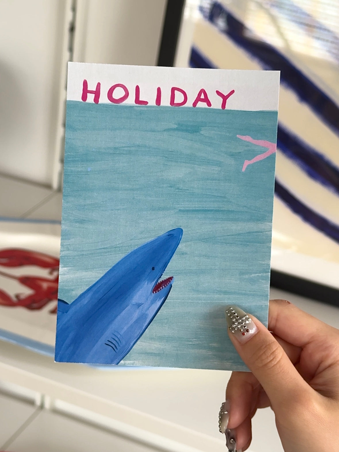 A hand with decorated nails holds a postcard featuring a shark, pink airplane, and "HOLIDAY"—a playful scene perfect for gifting or pairing with the Stacked Store Holiday Gift Card (Physical).