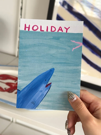 A hand with decorated nails holds a postcard featuring a shark, pink airplane, and "HOLIDAY"—a playful scene perfect for gifting or pairing with the Stacked Store Holiday Gift Card (Physical).