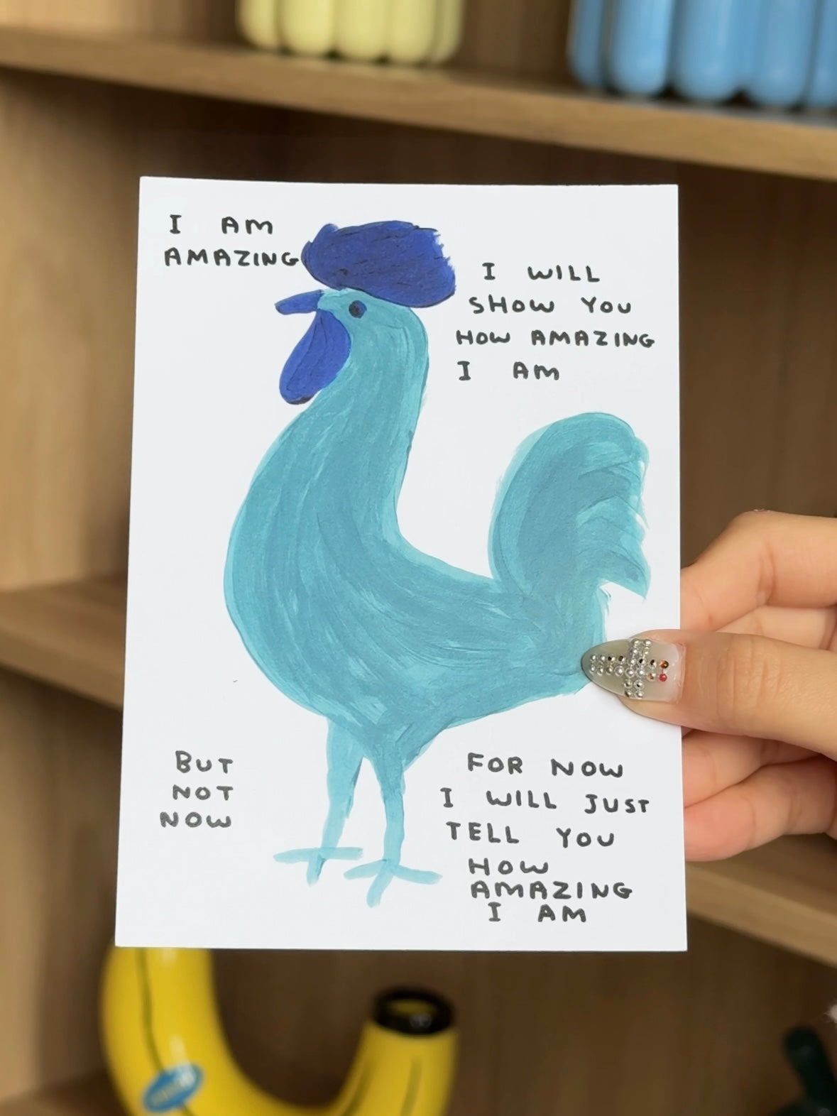A hand holds the Stacked Store "I Am Amazing Gift Card (Physical)" featuring a blue rooster and handwritten text: "I am amazing. I will show you how amazing I am. But not now. For now I will just tell you how amazing I am.