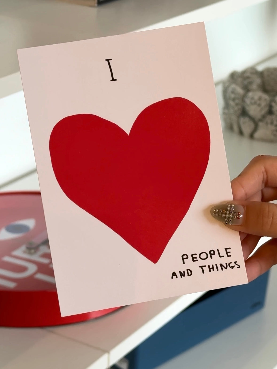 A hand holds the Stacked Store's I Love People Gift Card (Physical), featuring a bold red heart and the phrase "I love people and things"—a perfect choice for holiday gifting.