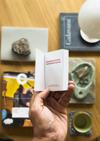 A hand holds the Stacked Store Object Porn Tiny Book by Stacked Store, open to a page reading "CENSORED FOR PRINT, UNCENSORED ONLINE" above a wooden table with a rock, books, a magazine, and a cup—available at Stacked Store.