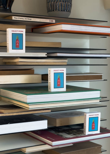 A modern bookshelf holds several Stacked Store Object Porn Tiny Books by Stacked Store, featuring a red bottle cover, stacked both vertically and horizontally—an ideal display for any contemporary space.
