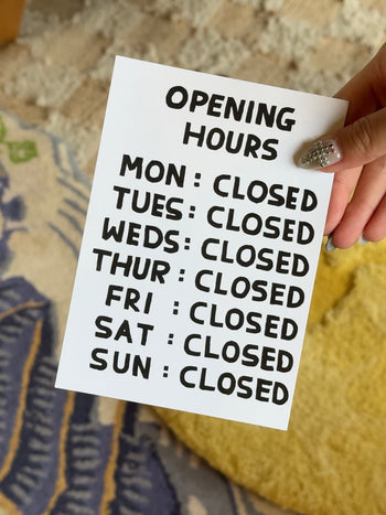 A hand holds the "Opening Hours Gift Card (Physical)" by Stacked Store, with all days marked CLOSED—quirky David Shrigley style against a patterned rug and bright yellow accent. A unique holiday gift idea!.