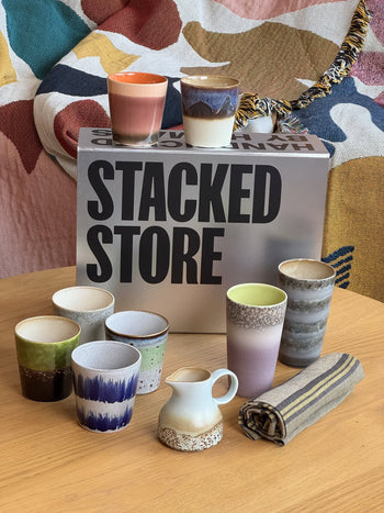 The Stacked Store The Coffee Lover Gift Box features colorful ceramic cups, a small pitcher, and a striped cloth on a wooden table—an ideal housewarming gift for new homeowners. A patterned blanket completes the cozy scene.