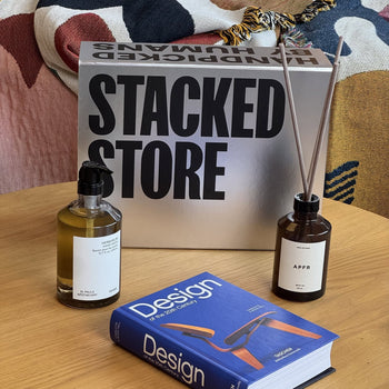 A silver Stacked Store The Design Enthusiast Gift Box sits on a wooden table with room fragrance, a reed diffuser, and a blue design coffee table book against a colorful patterned fabric backdrop. Brand: Stacked Store.