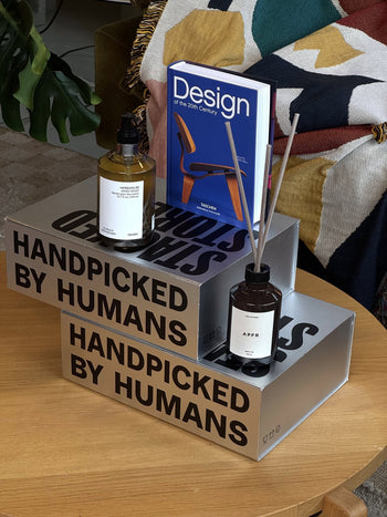 The Stacked Store The Design Enthusiast Gift Box features stacked boxes with a fragrance bottle, reed diffuser, and the book "Design of the 20th Century"—a unique gift for design lovers. A blanket and plant create a cozy background.