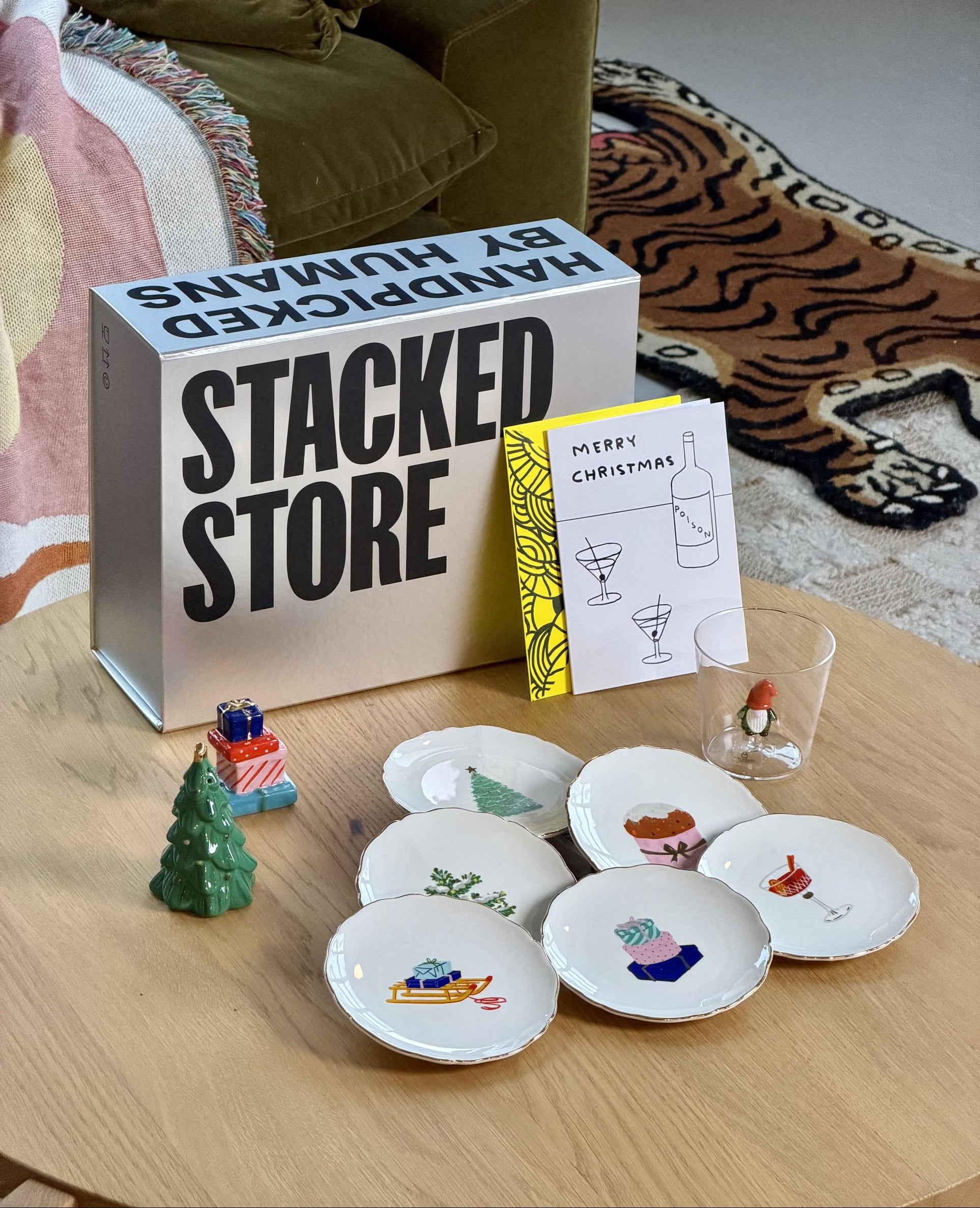 An illustrated food-themed Christmas tableware set, glass with figurine, ceramic tree, and matchbox are arranged next to Stacked Store's Festive Holiday Gift Box and Merry Christmas card. A sofa and rug are visible in the background.