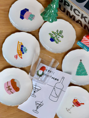 The Festive Holiday Gift Box by Stacked Store includes seven small Christmas plates with festive designs and a Merry Christmas card featuring a wine bottle and two figures—ideal for gifting or same-day delivery, beautifully arranged on a wooden surface.