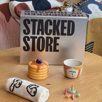 The Stacked Store The Homebody Gift Box by Stacked Store rests on a wooden table, featuring a pancake-shaped mug, a face-illustrated cup, rolled towel, colorful small cones, and a patterned blanket in the background.