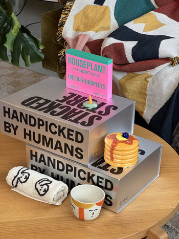 A cozy scene features Stacked Store The Homebody Gift Box: two silver HANDPICKED BY HUMANS boxes, a towel and cup with faces, ceramic pancake plates, and a pink incense box—an ideal luxury home gift from Stacked Store.