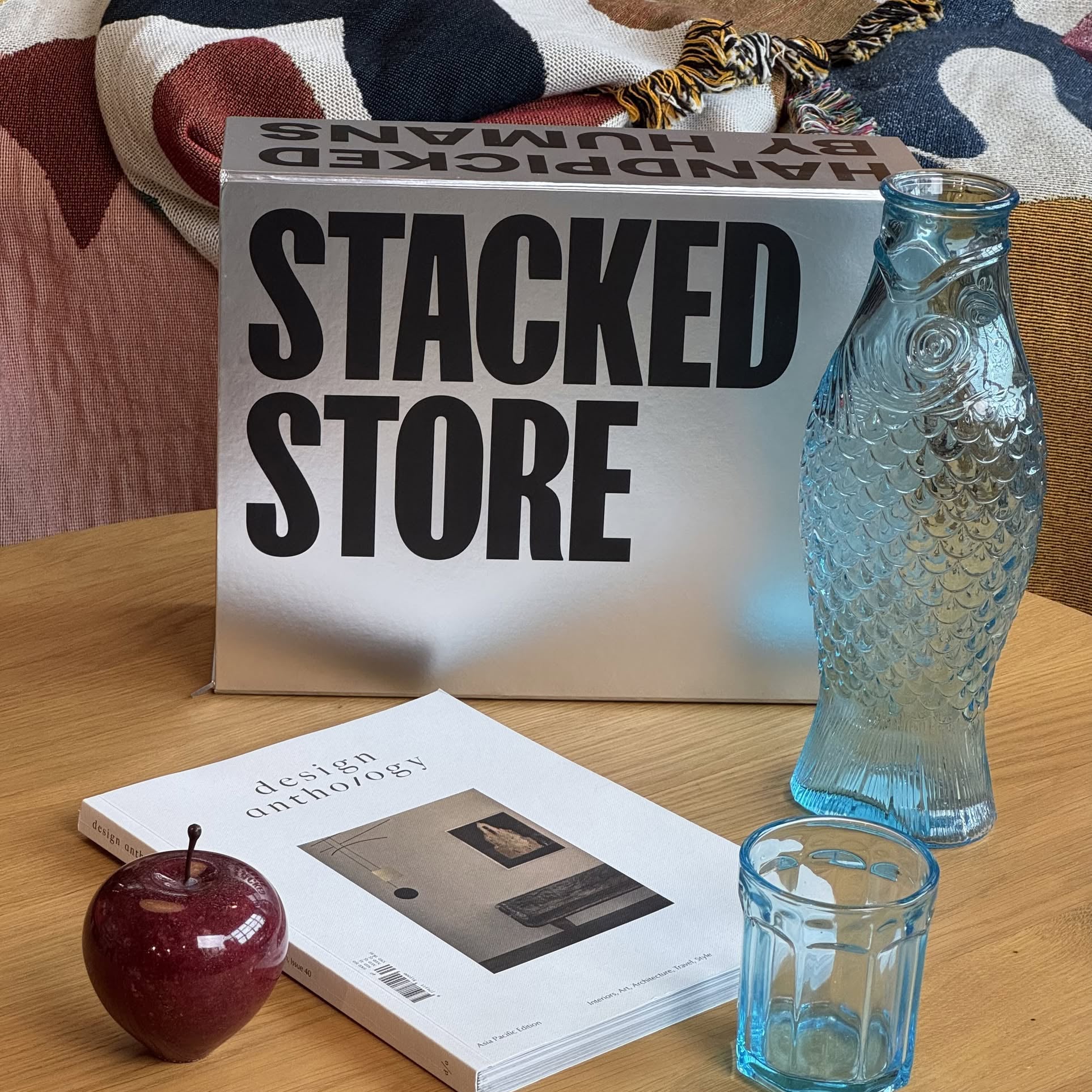 A table with a red apple, glass cup, blue fish-shaped bottle, magazine, and Stacked Store The Minimalists Gift Box—ideal for coffee lovers—set against a colorful fabric backdrop.