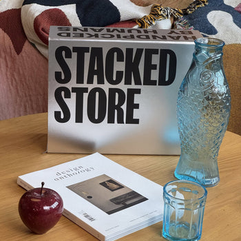 A table with a red apple, glass cup, blue fish-shaped bottle, magazine, and Stacked Store The Minimalists Gift Box—ideal for coffee lovers—set against a colorful fabric backdrop.