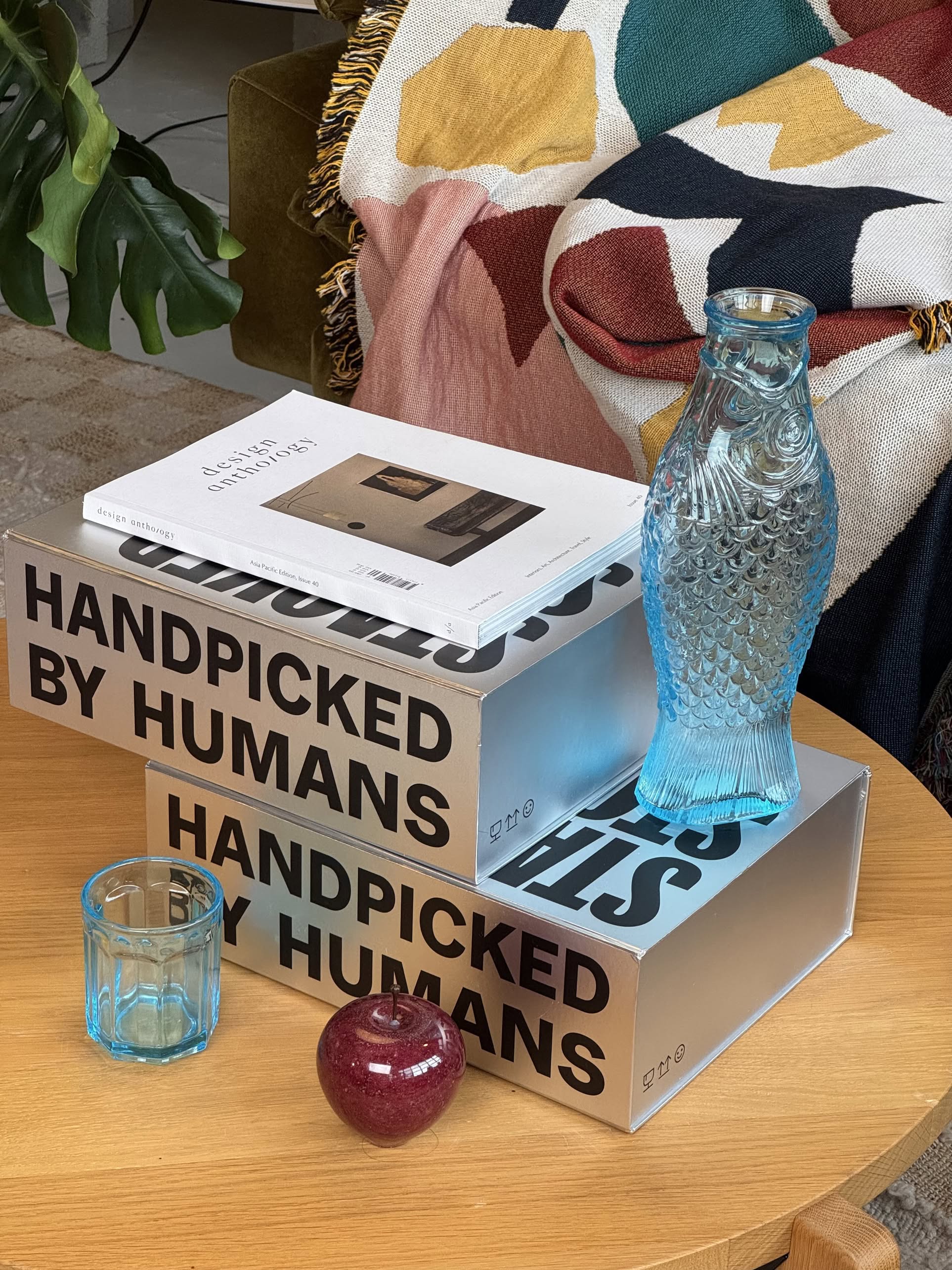 A wooden table displays the Stacked Store The Minimalists Gift Box, featuring two stacked books, a blue fish-shaped vase, a small blue glass, a red ornament, and a colorful blanket draped over a green couch in the background.