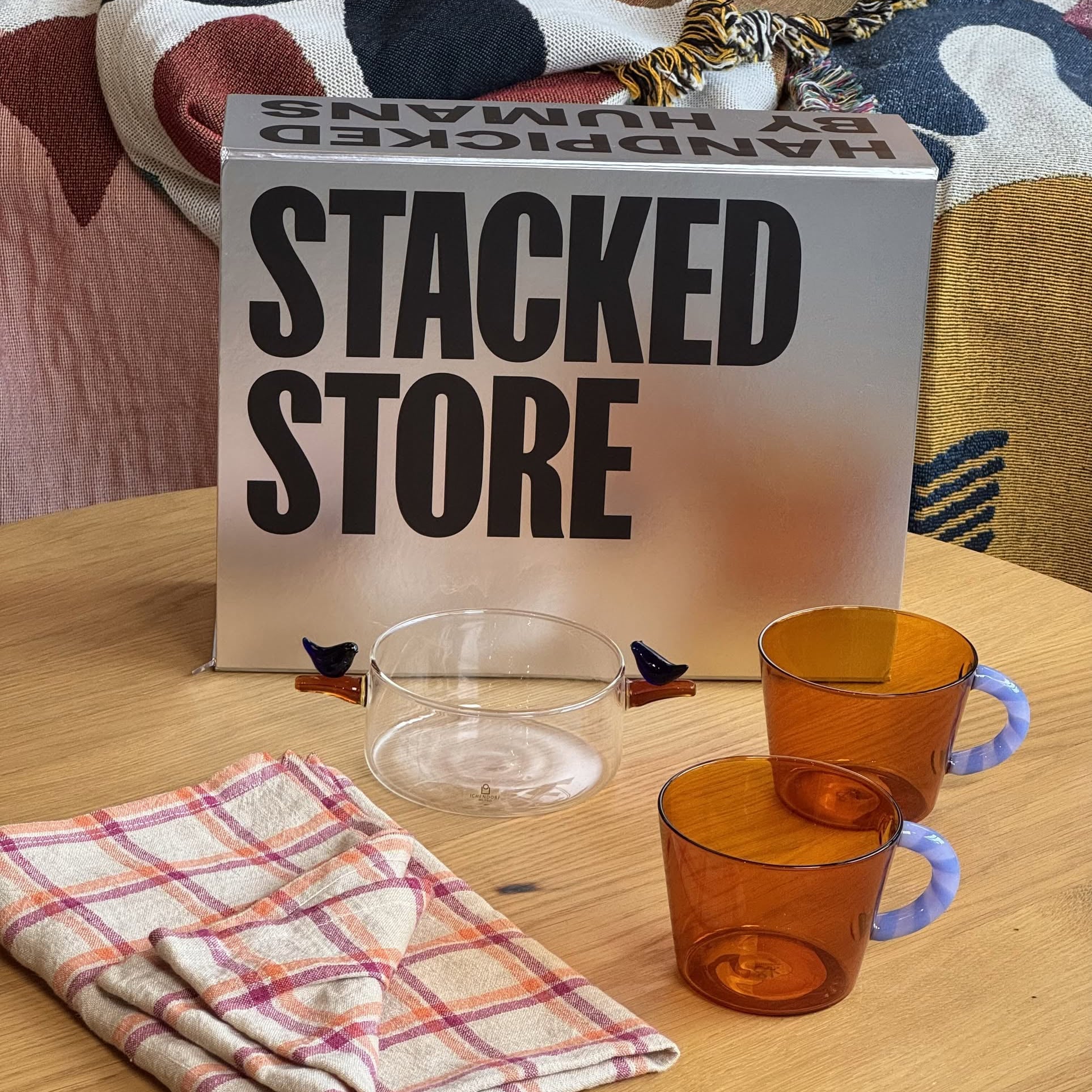 The Stacked Store The Modern Host Gift Box includes a silver box, two amber mugs, a clear glass bowl, two checkered cloths, and a colorful blanket—the ideal gift for design-forward minimalists.