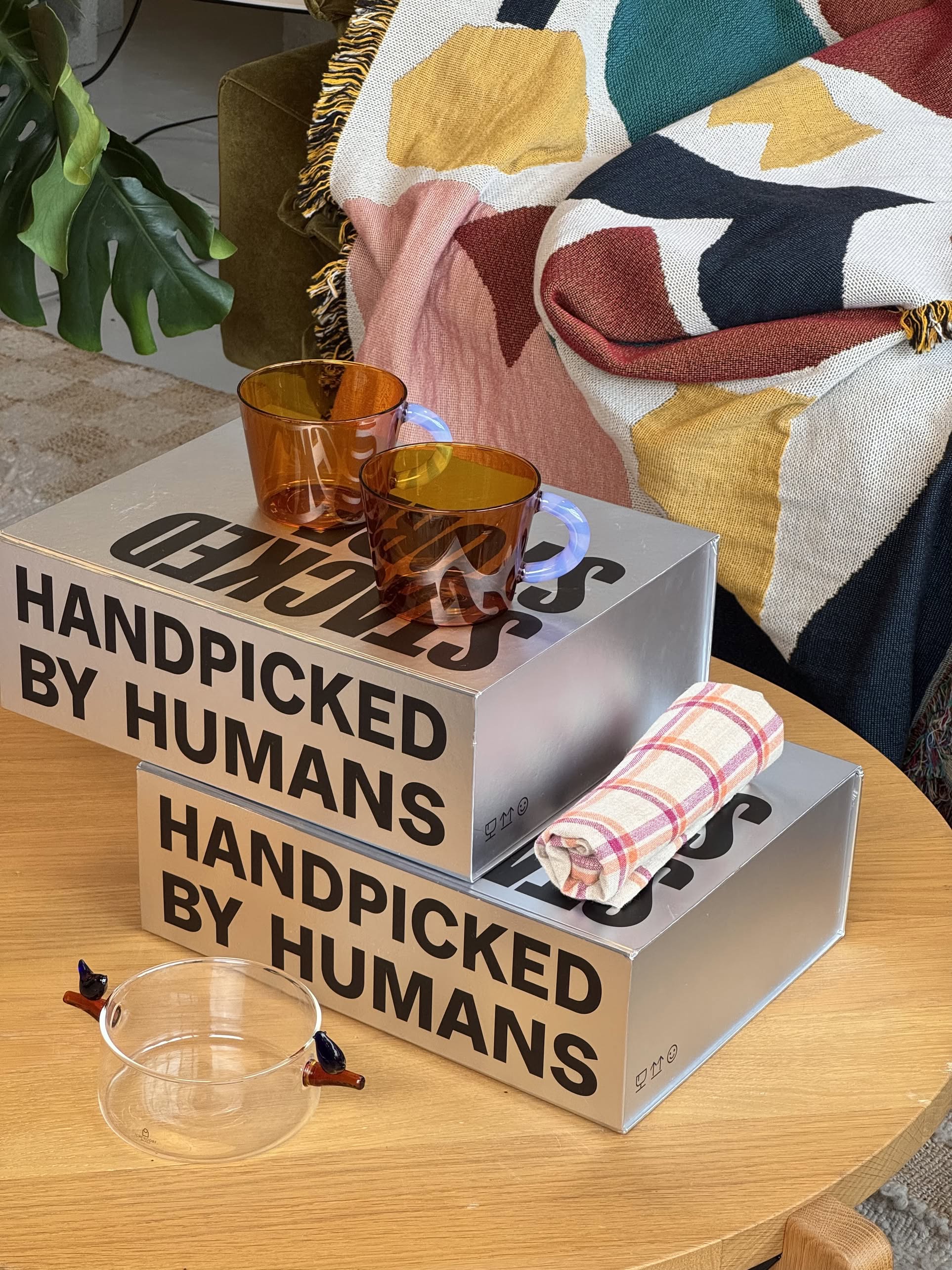Two amber glass cups and a checkered cloth rest on stacked boxes labeled "Handpicked by Humans." The Stacked Store The Modern Host Gift Box is a chic, modern gift set—complete with a patterned blanket over a green chair.